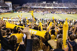 Crowd at a packed Appalachian State football game waving yellow spirit sticks beneath stadium lights.